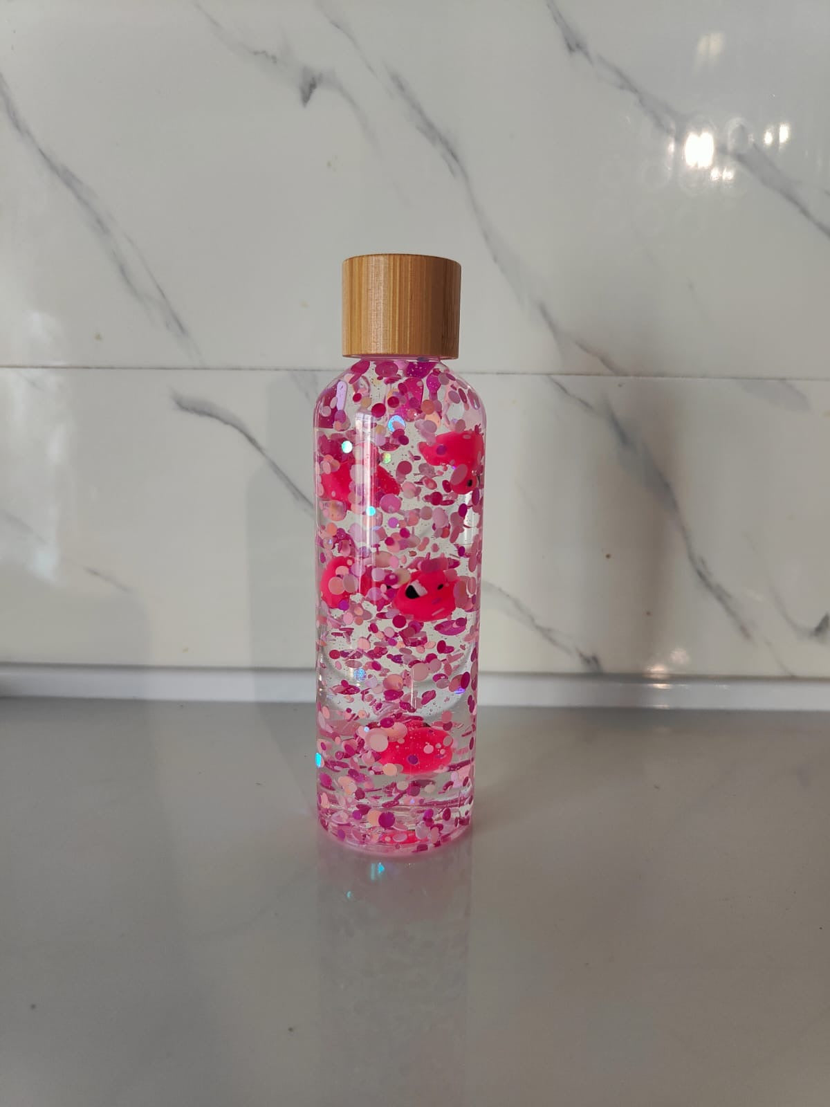 Bouteille sensorielle avec des flamants roses qui flottent parmi des paillettes roses. Effet visuel doux, fluide et apaisant. Créée à la main en France