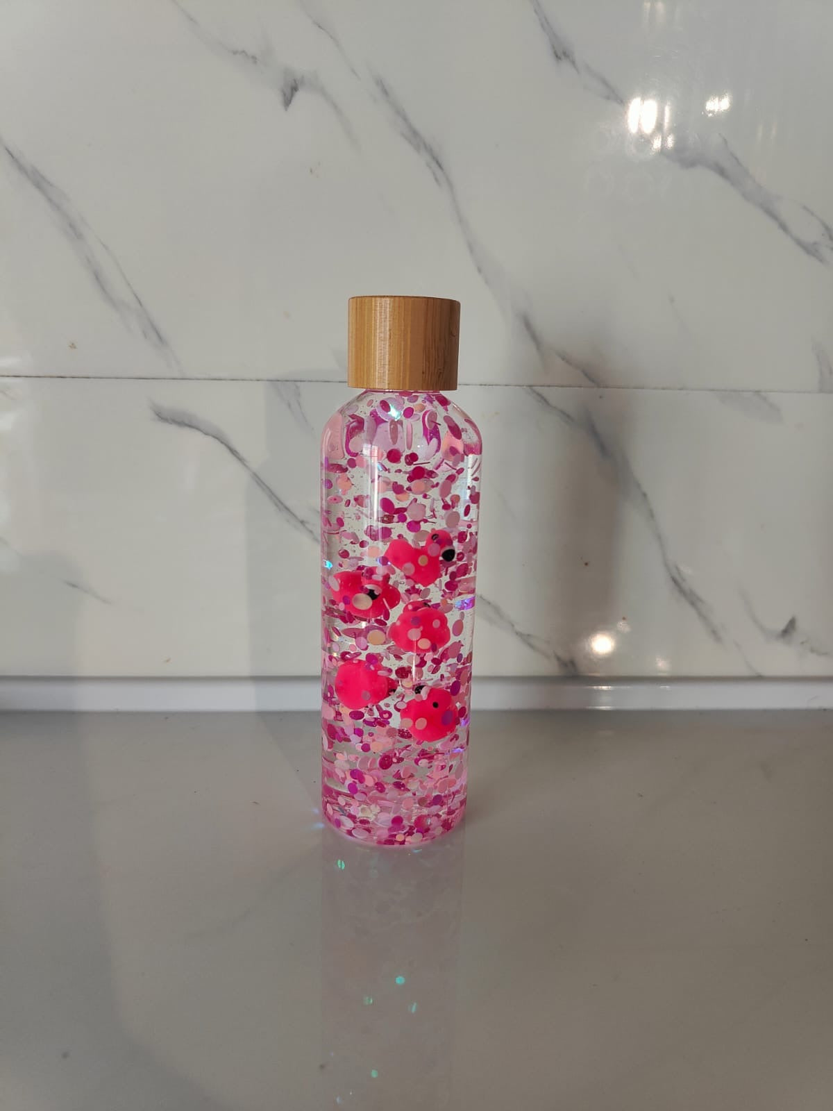 Bouteille sensorielle avec des flamants roses qui flottent parmi des paillettes roses. Effet visuel doux, fluide et apaisant. Créée à la main en France