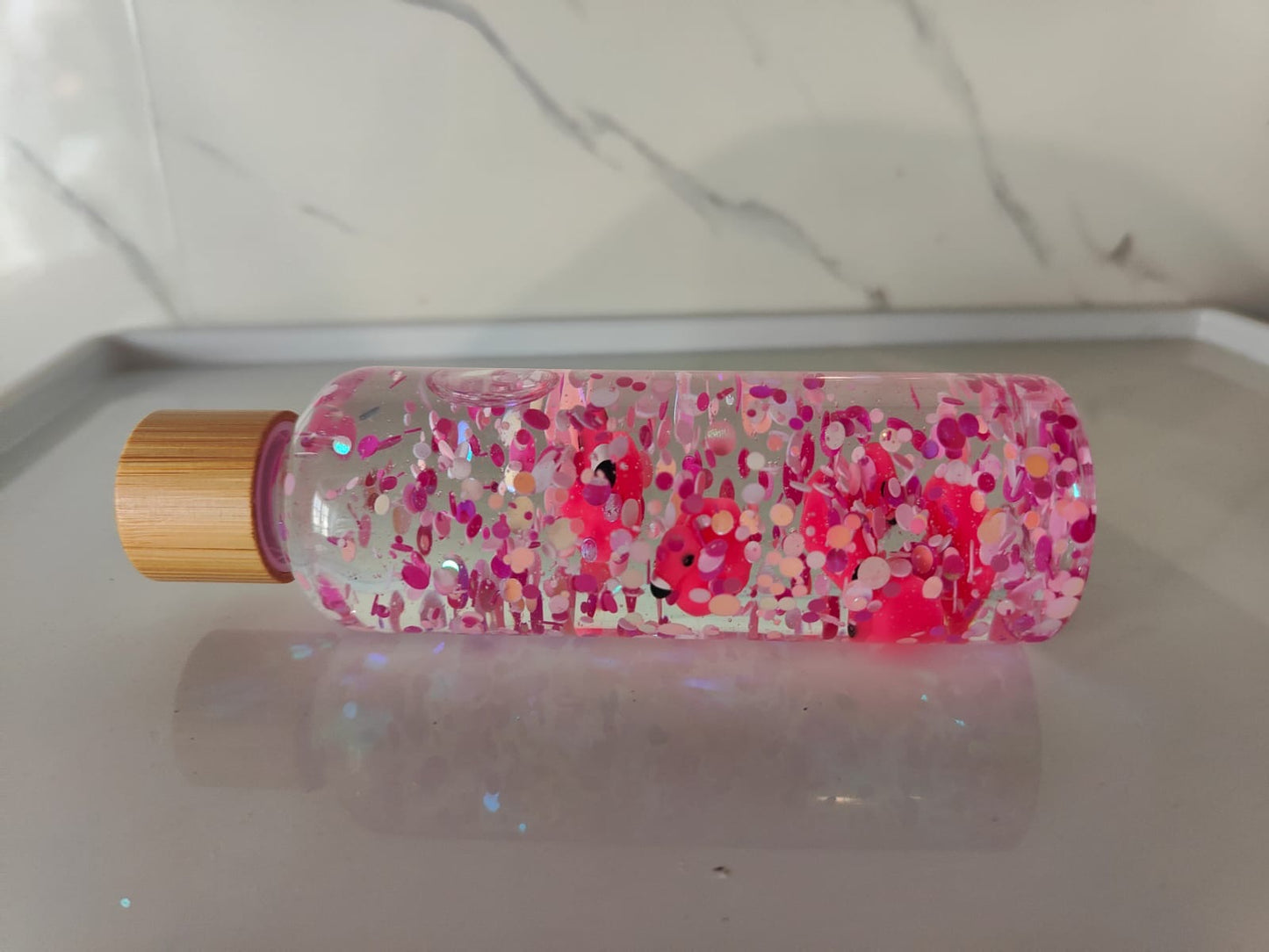 Bouteille sensorielle avec des flamants roses qui flottent parmi des paillettes roses. Effet visuel doux, fluide et apaisant. Créée à la main en France