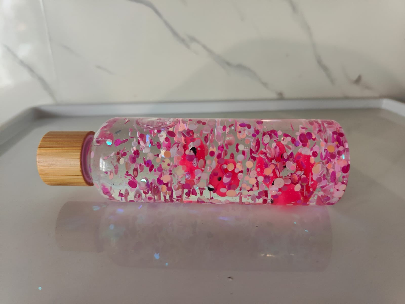 Bouteille sensorielle avec des flamants roses qui flottent parmi des paillettes roses. Effet visuel doux, fluide et apaisant. Créée à la main en France